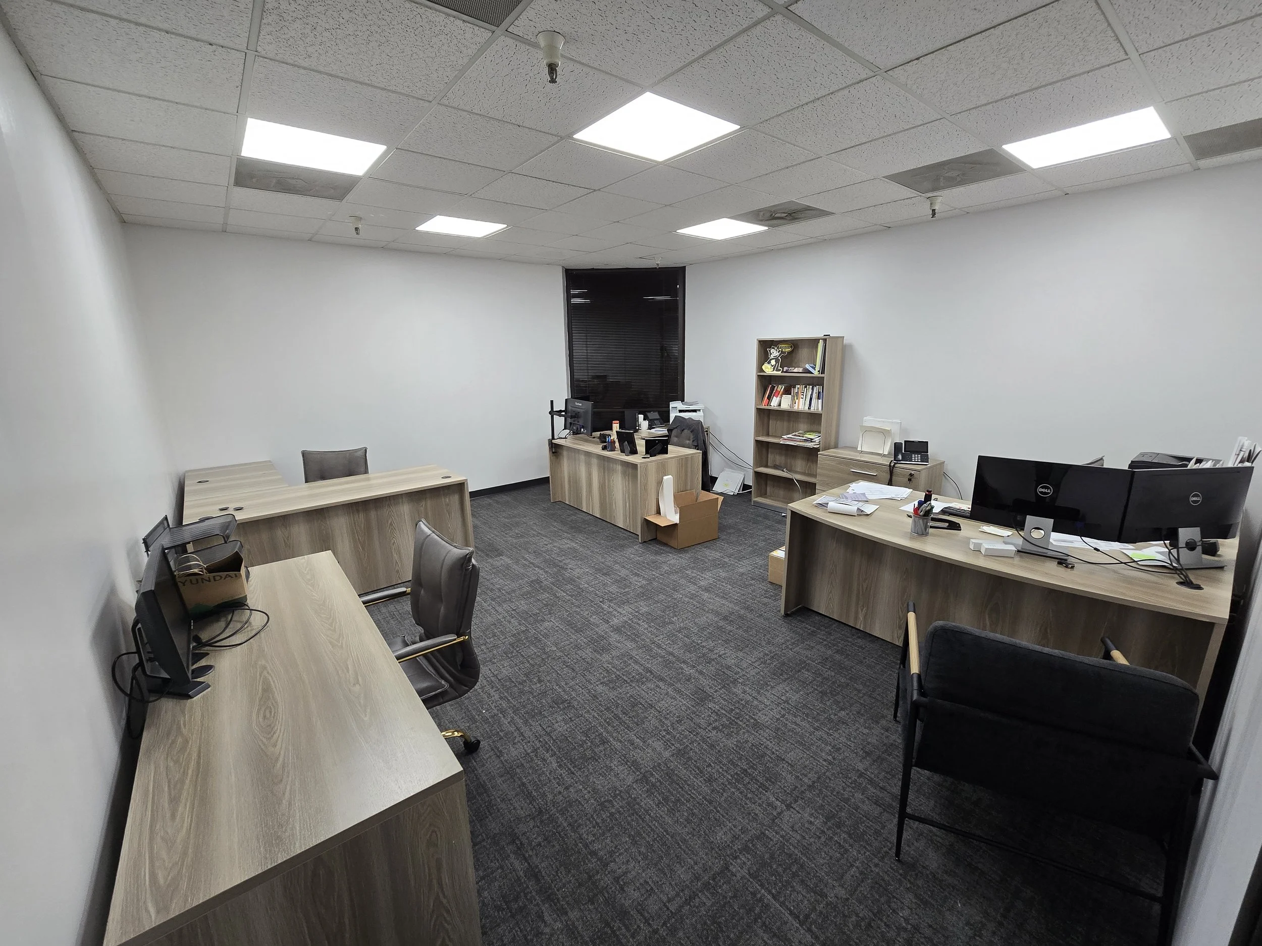 Empty office with three desks, two monitors, office chairs, bookshelf, and scattered papers.