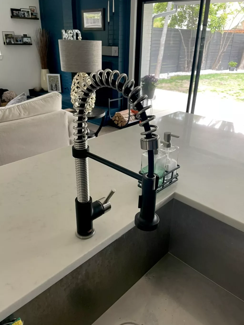 Close-up of a modern kitchen faucet with a flexible, coiled spring design, installed on a white countertop next to a window and a soap dispenser.
