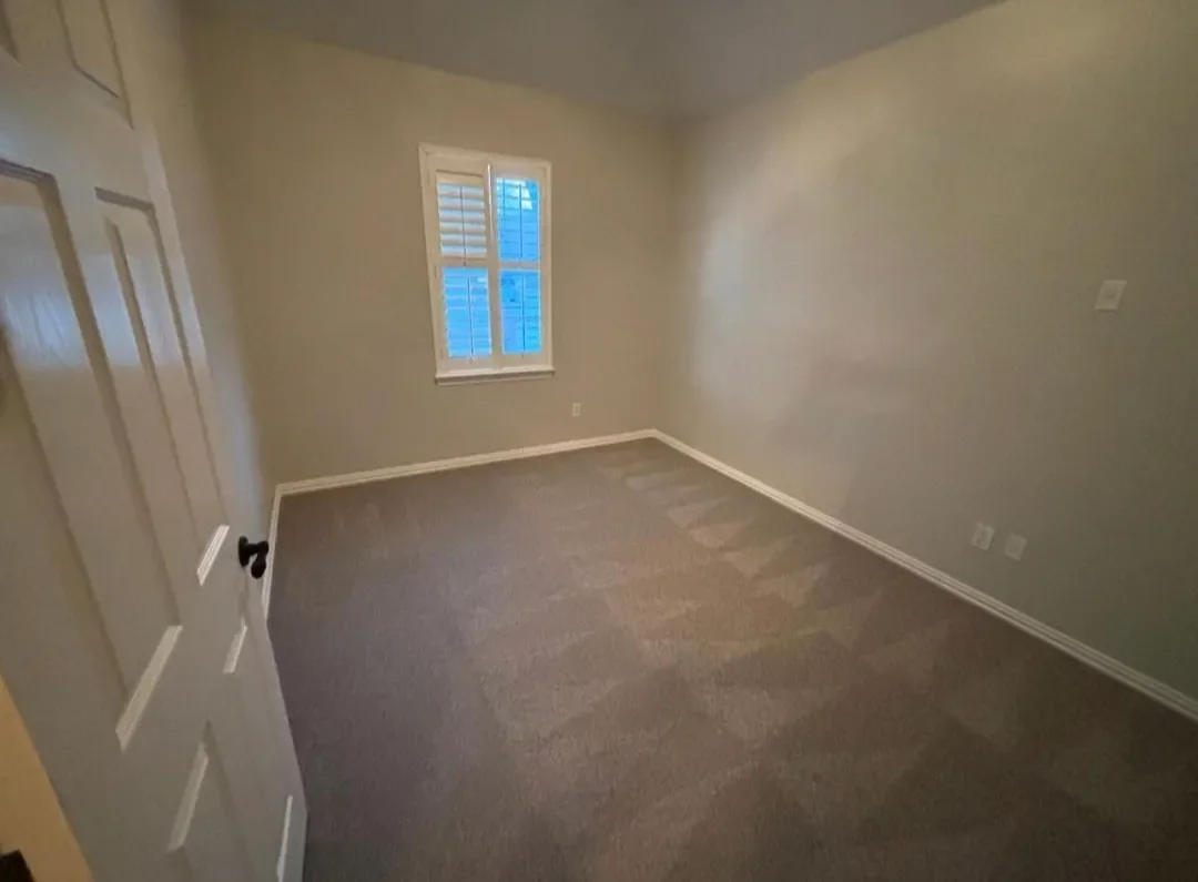 Empty bedroom with beige walls, grey carpet, a window with blue outside view, and a closed door.