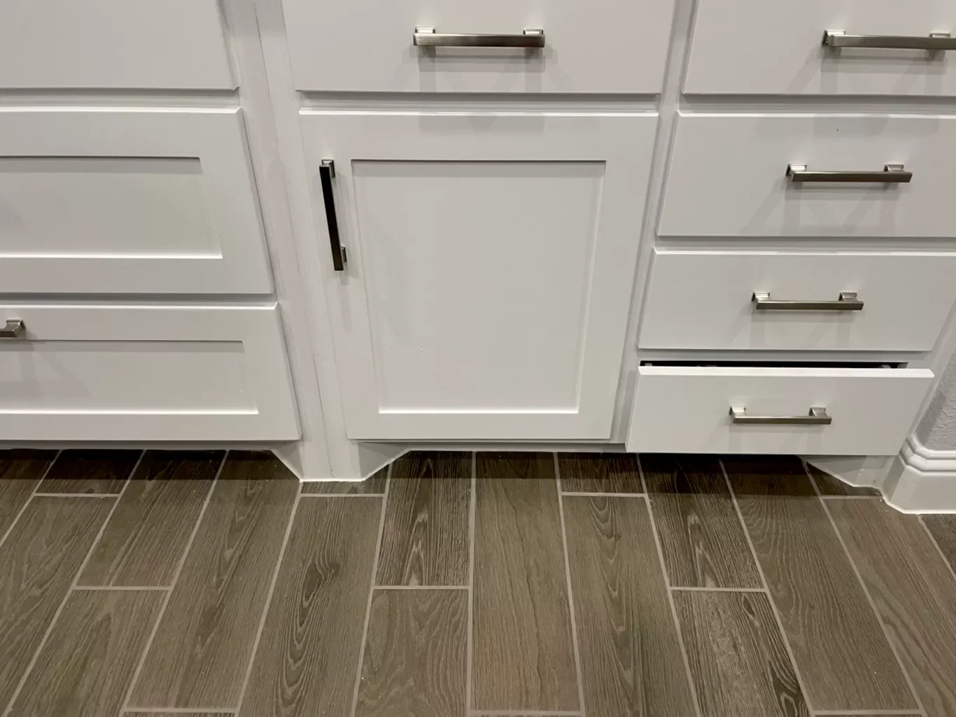 Close-up of white kitchen cabinets with silver handles and a tiled floor, showing one cabinet drawer partially open.