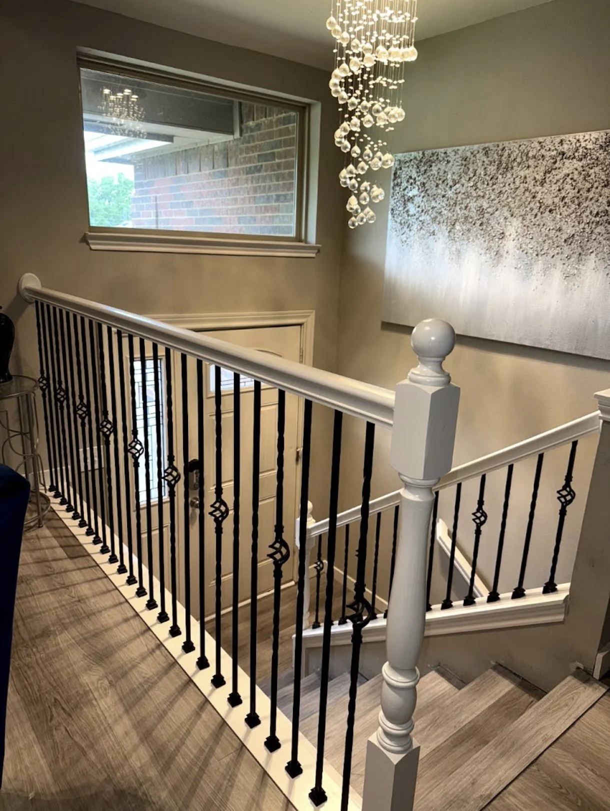Indoor staircase with wooden flooring, black iron balusters, white handrail, and decorative newel post, near a window and modern artwork on the wall.