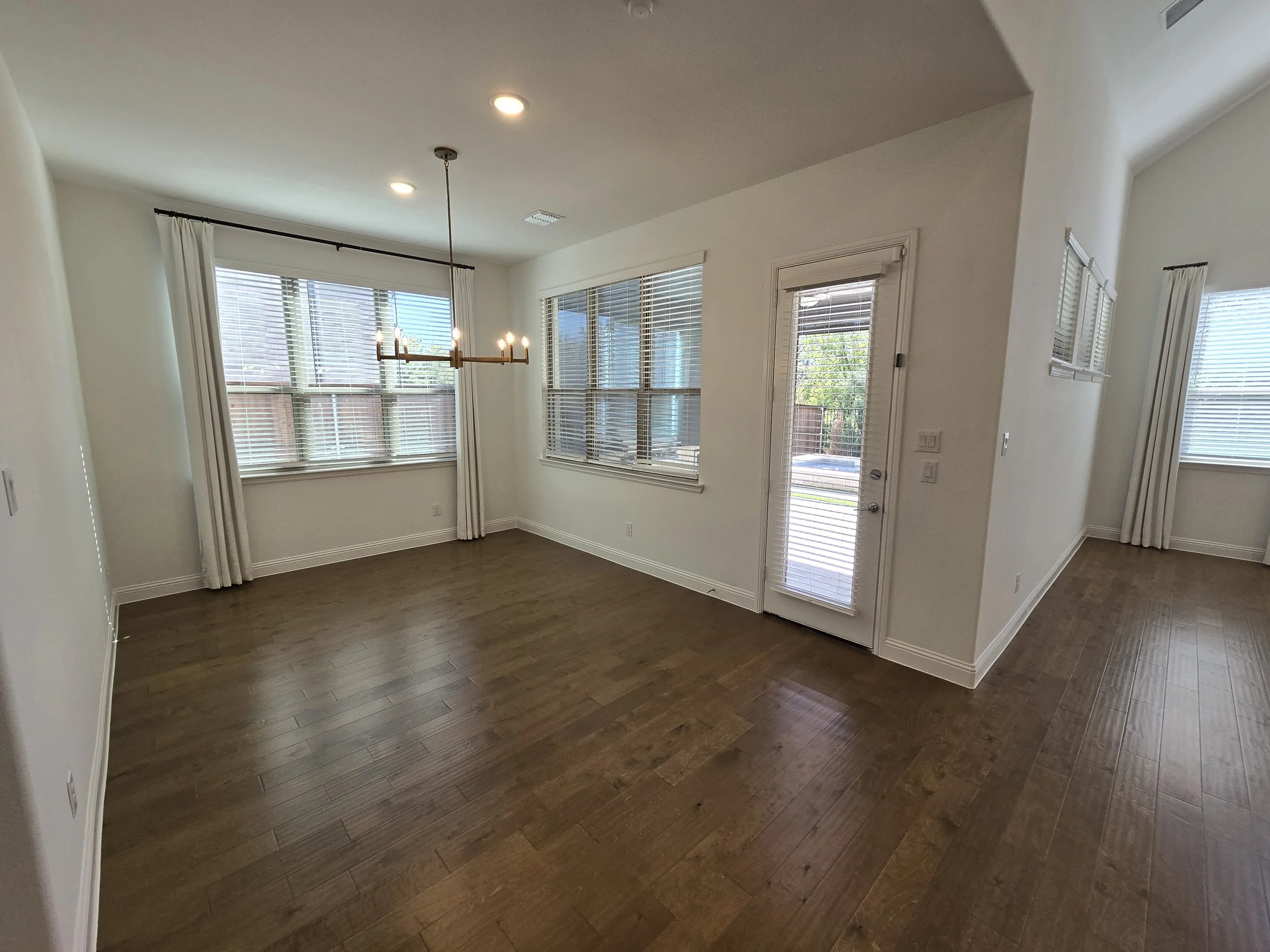 Empty room with wooden flooring, white walls, studded with large windows with white blinds and curtains, a glass door with blinds, and a chandelier hanging from the ceiling.