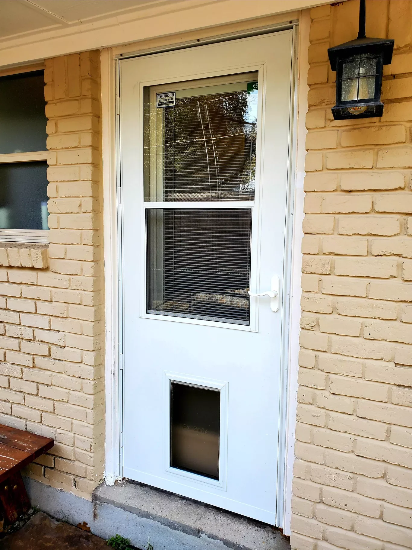 White storm door with a window and a pet door installed in a brick house exterior with a black outdoor light fixture.