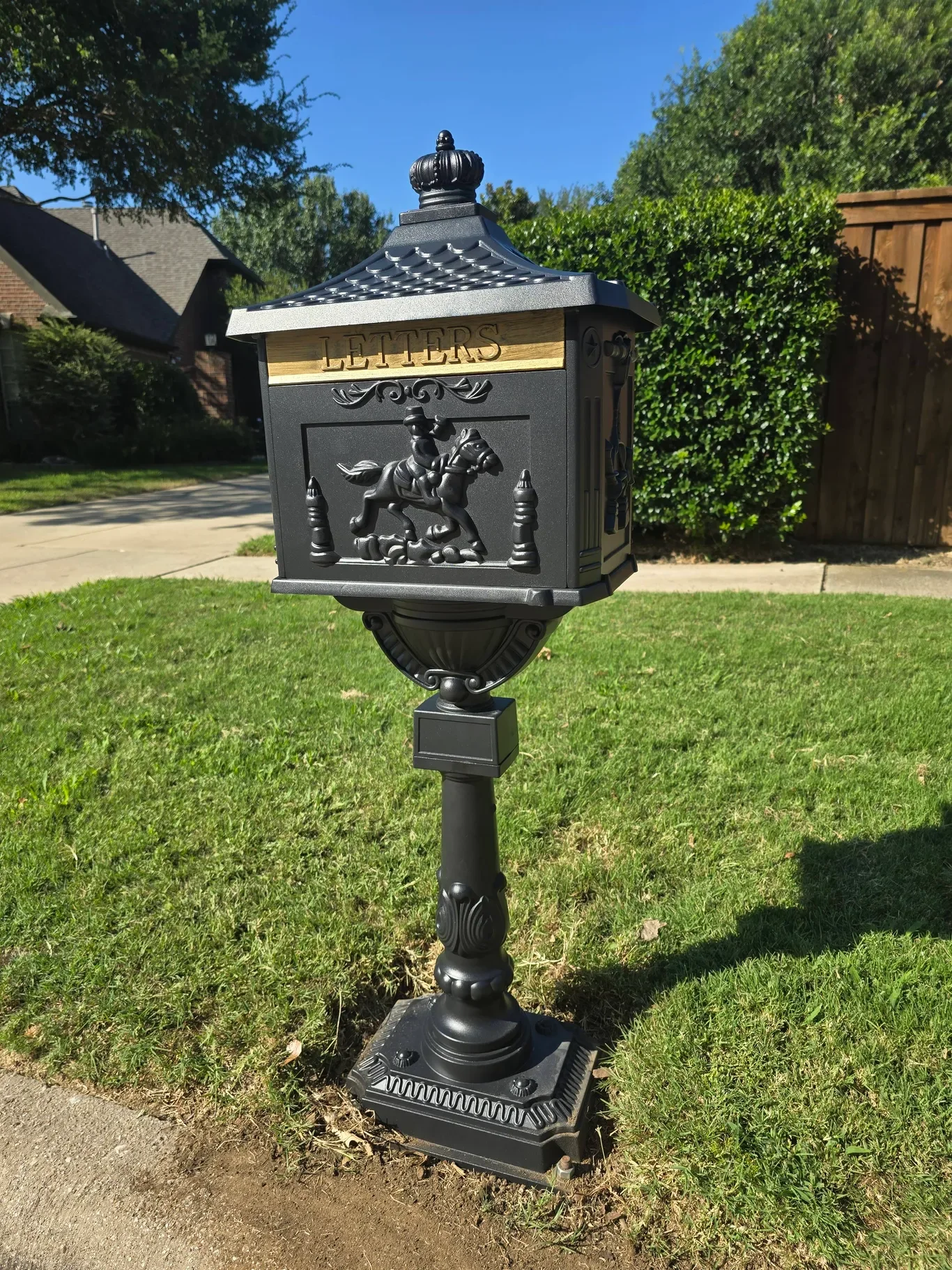 A black metal mailbox with a wooden panel labeled 'LETTERS' at the top, featuring a horse and rider design, mounted on a decorative post in a grassy yard.