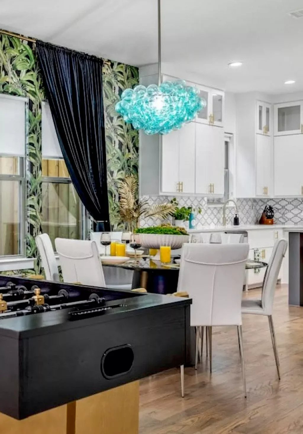 Open kitchen and dining area with white cabinets, patterned backsplash, a black and gold foosball table, white chairs around a dining table set with glasses of orange juice and glasses of red wine, and a blue-green chandelier hanging from the ceiling