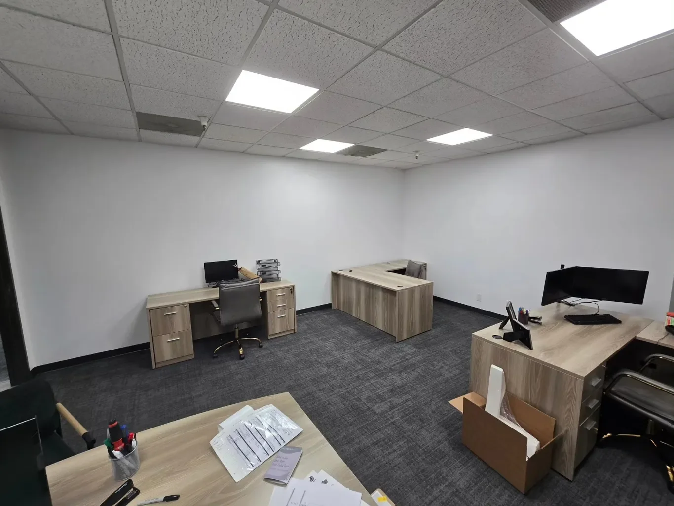 Empty office room with three desks, two of which have computer monitors and office supplies. Gray carpet, white walls, and a dropped ceiling with fluorescent lights.