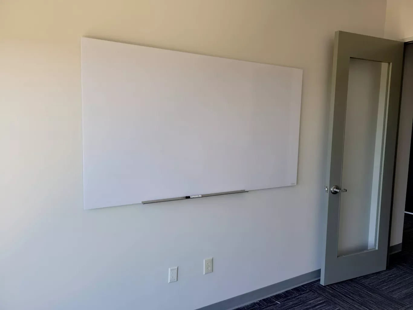 Empty whiteboard on a wall in a room with two electrical outlets and a glass-paneled gray door.