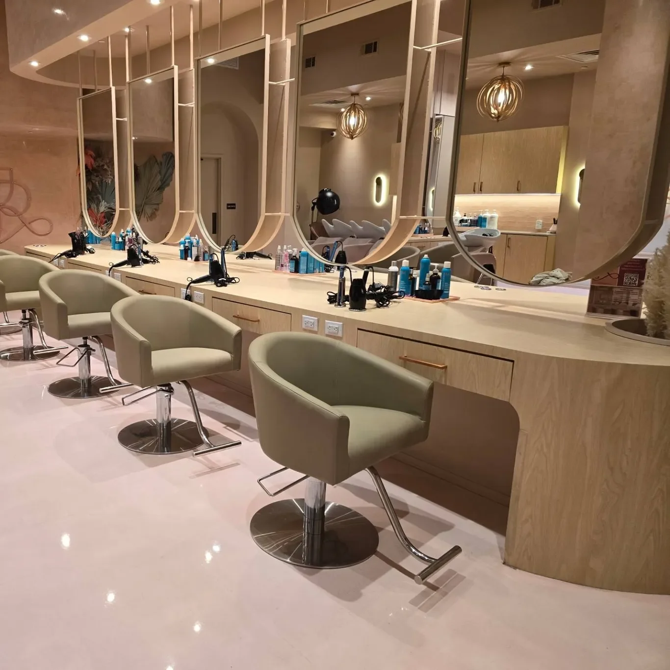 A modern beauty salon or hairdressing station with beige chairs, large mirrors, hairdryers, and hair products on the counter, with warm lighting and contemporary decor.
