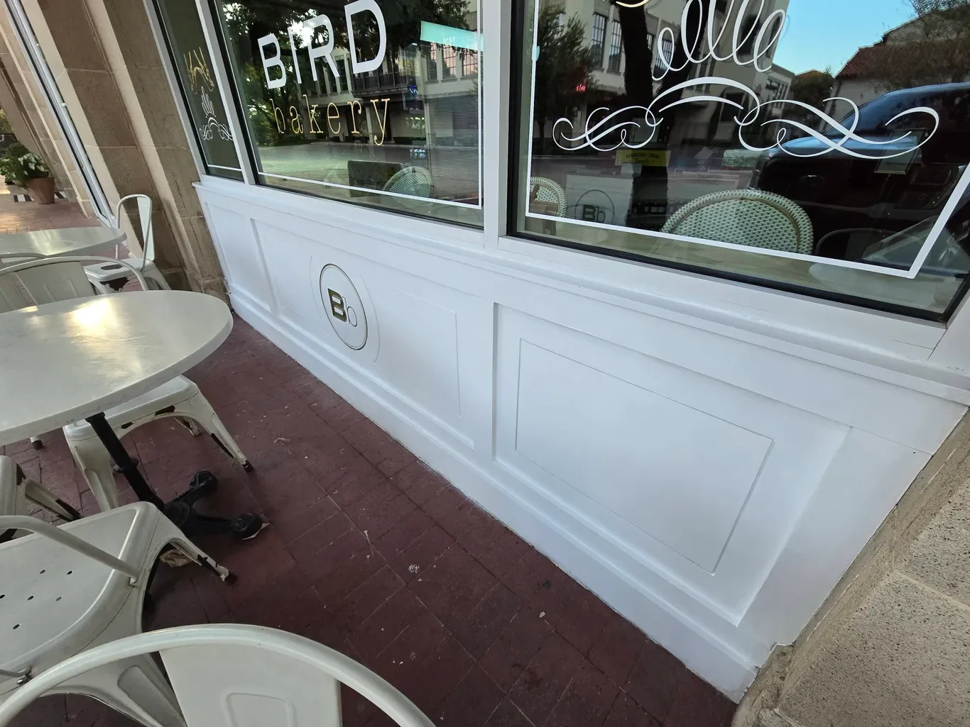 Exterior view of a bakery storefront with white window frames and outdoor seating area, including white chairs and tables, and glass windows with bakery signage.
