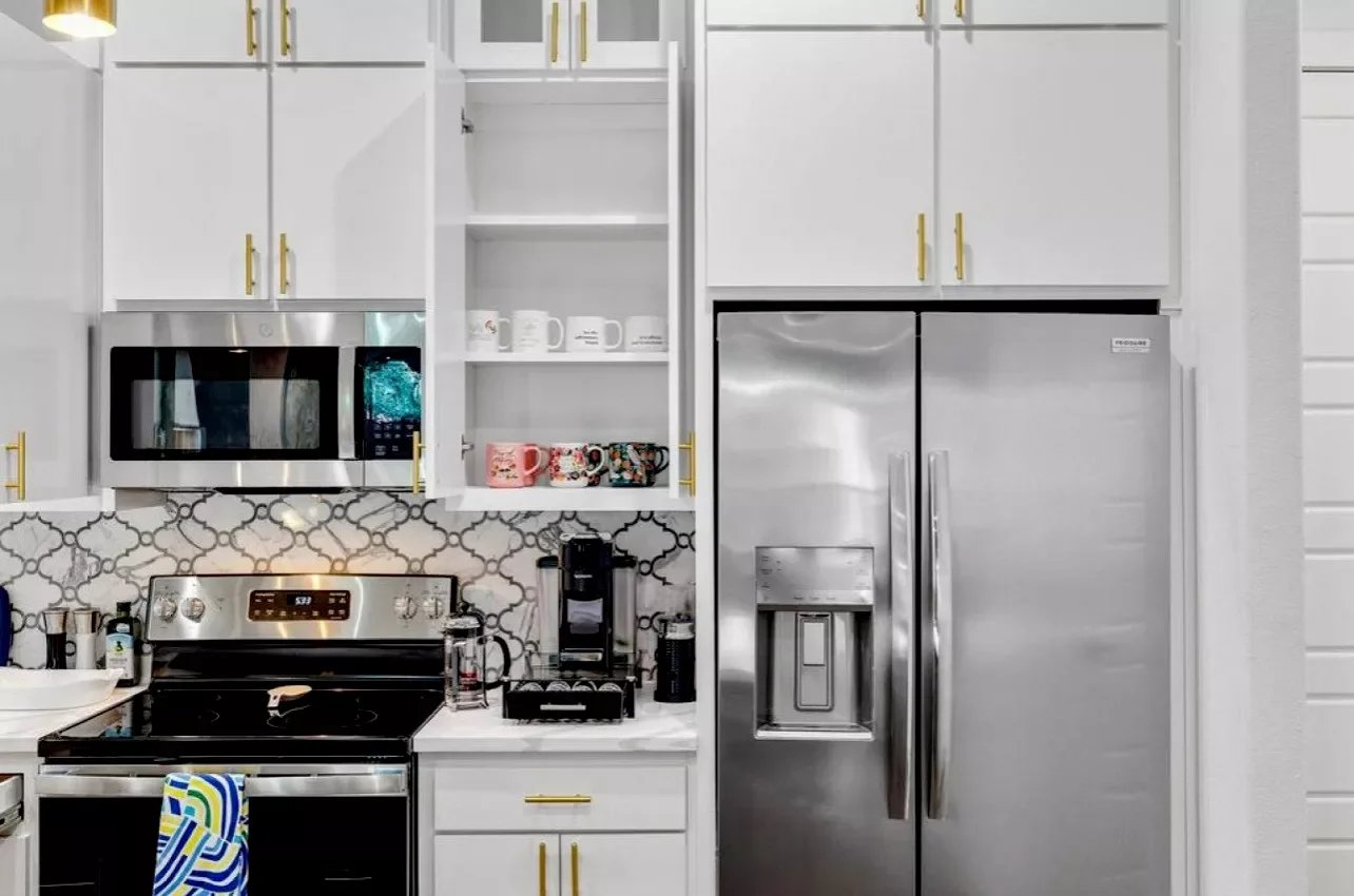 Kitchen with white cabinets, a stainless steel microwave, a double-door refrigerator, open shelves with mugs, a stove, and a coffee maker.