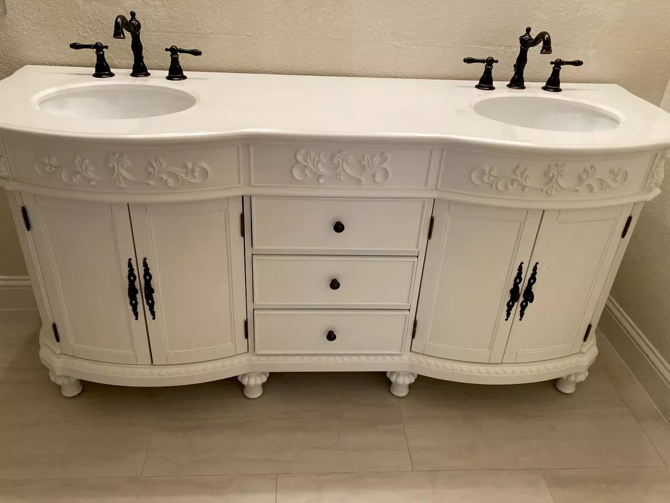 White vintage double-sink bathroom vanity with black fixtures, decorative carved details, and three drawers in the center, set against a beige wall.