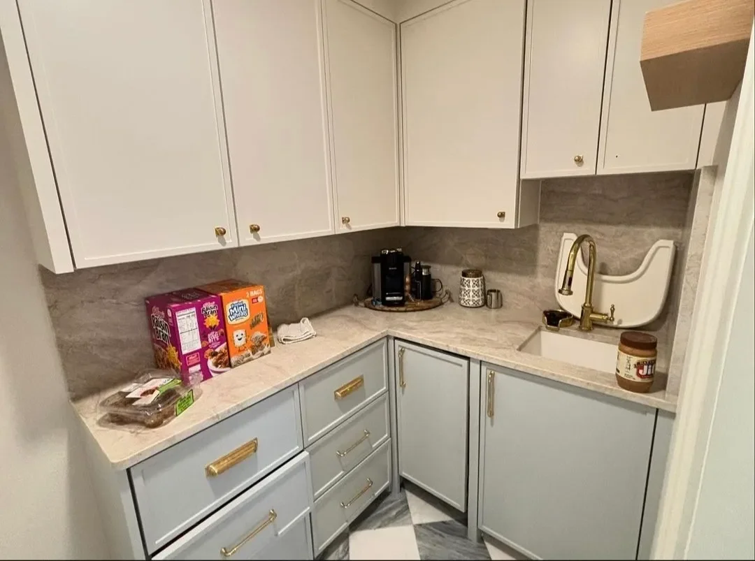 A small kitchen corner with white and light blue cabinets, Gold handles, a marble countertop, a small sink, a brass faucet, a coffee maker, cereal boxes, a jar of Jif peanut butter, and a few other small items.