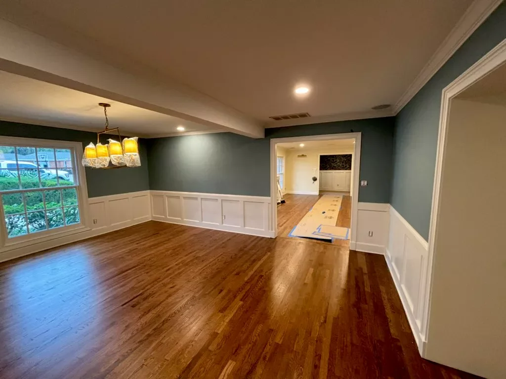 Empty dining room with hardwood floors, a window with a view of a yard, a chandelier, and wainscoting on the walls. A doorway leads to another room with similar flooring and some construction tape.