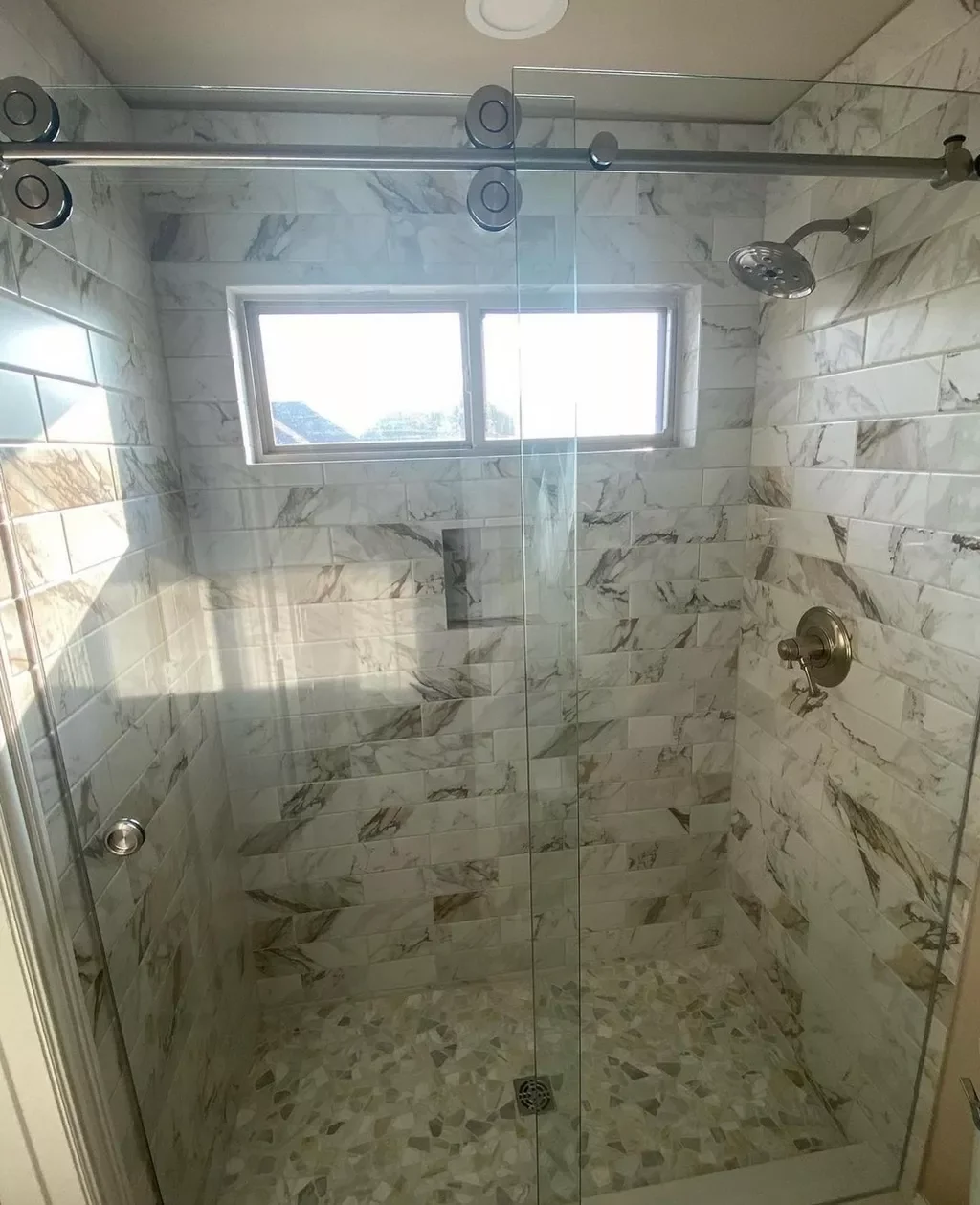 A walk-in shower with marble tile walls and a small window, enclosure with a glass door, a ceiling-mounted showerhead, and a brass control handle.
