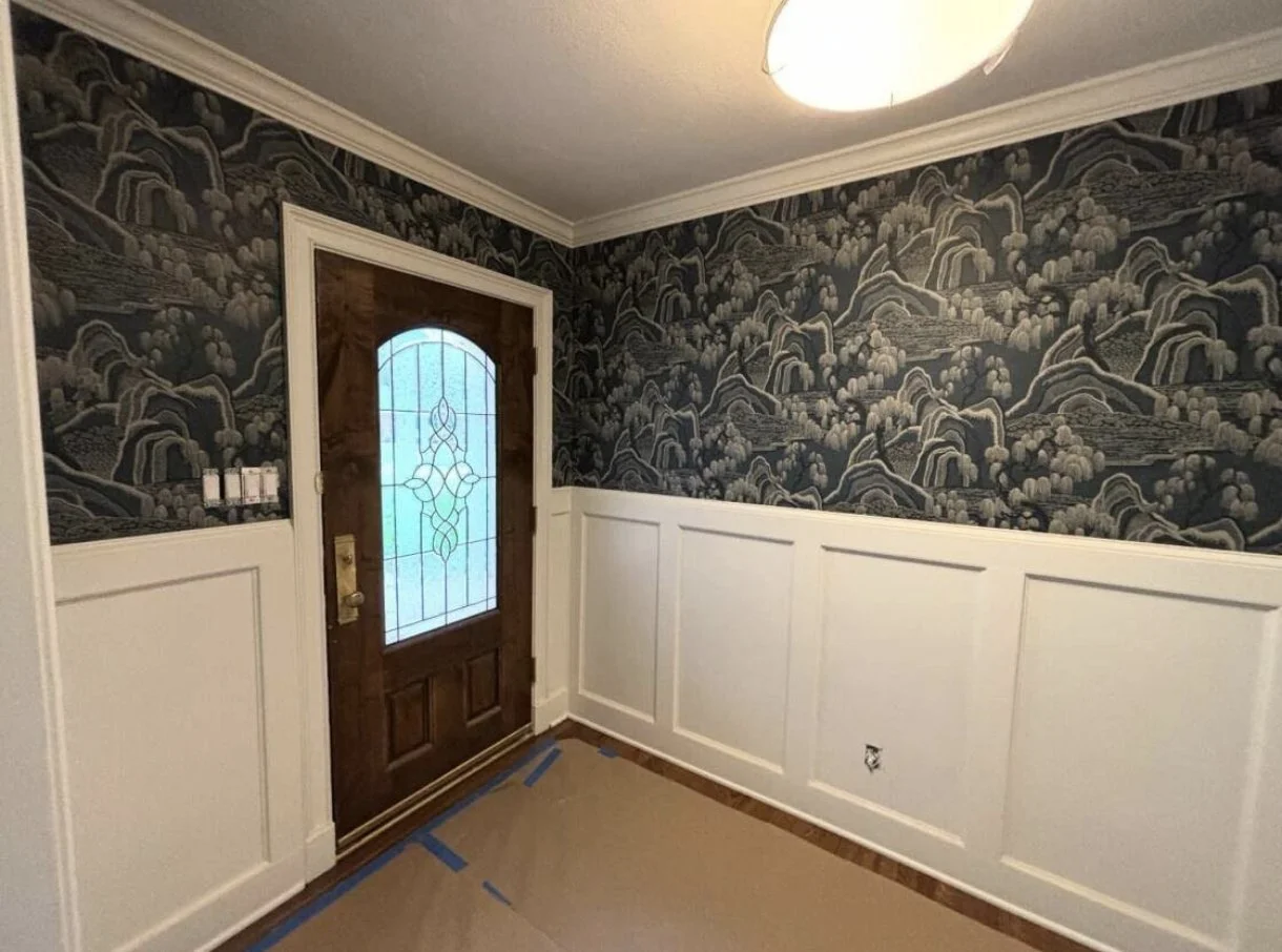 Interior view of a room with a wooden front door, decorative stained glass window, dark patterned wallpaper, and white wainscoting, with some painter's tape on the floor.