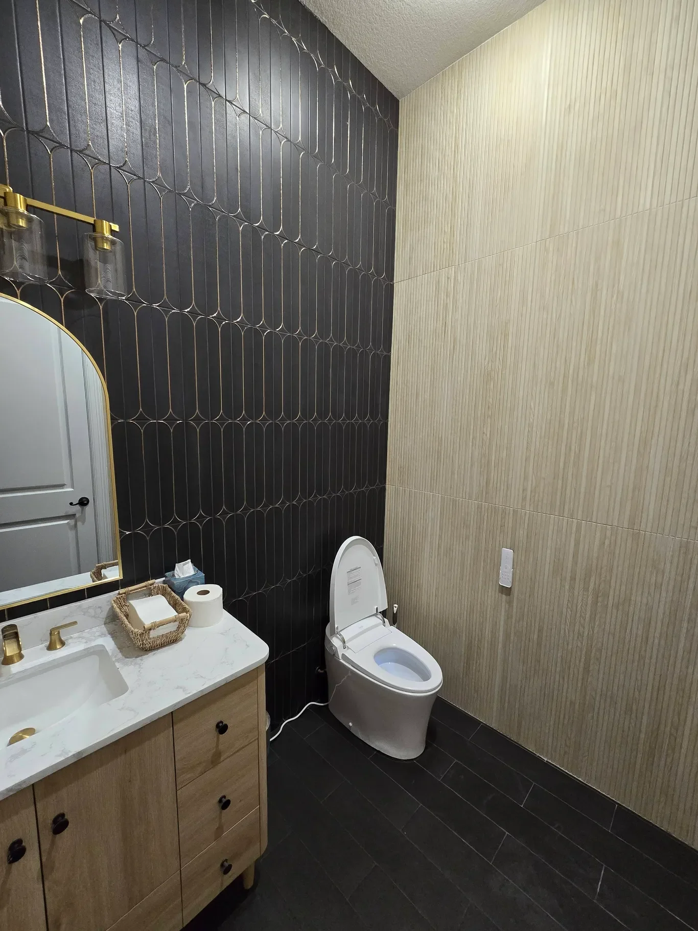 A modern bathroom with a black tiled accent wall, a white toilet, and a wooden vanity with a marble countertop and gold fixtures.