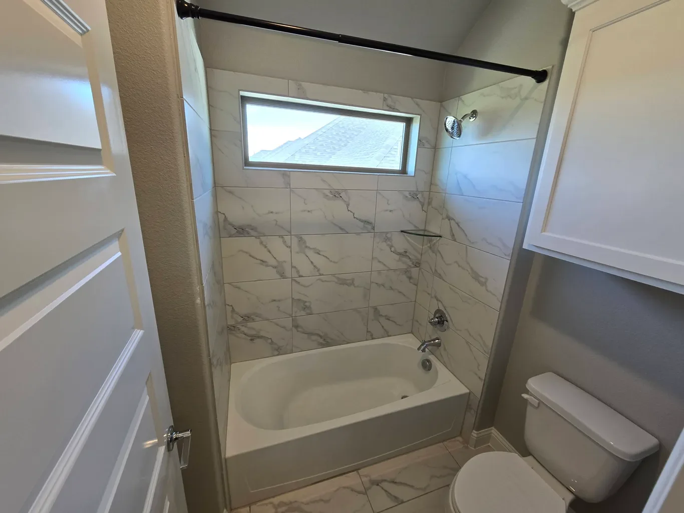 Bathroom with a bathtub, marble tiles, a small horizontal window above the tub, a showerhead, and a toilet in the corner.