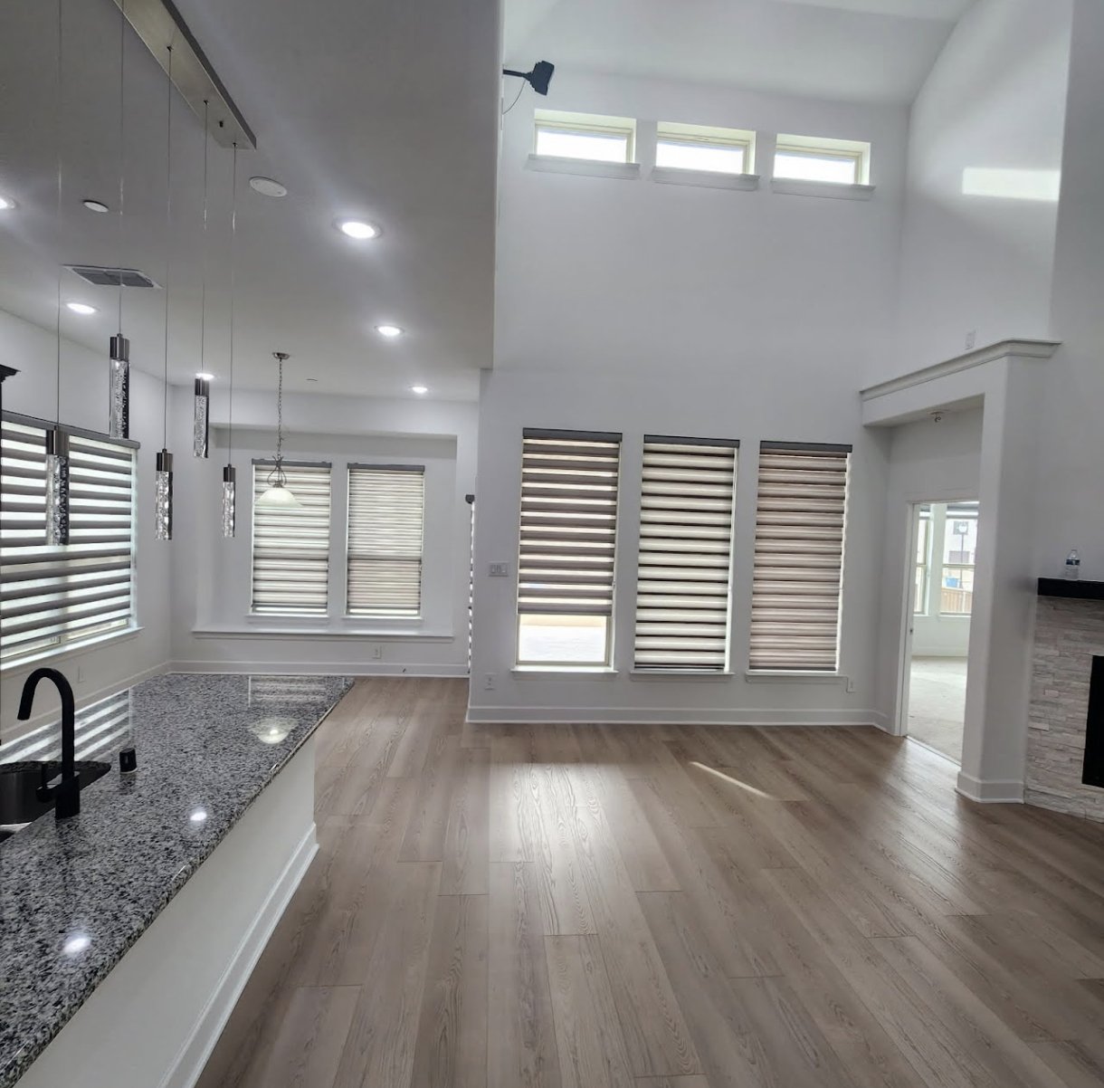 A spacious modern living room with large windows covered by zebra blinds, light wood flooring, and a partial view of a kitchen with a granite countertop and pendant lights.