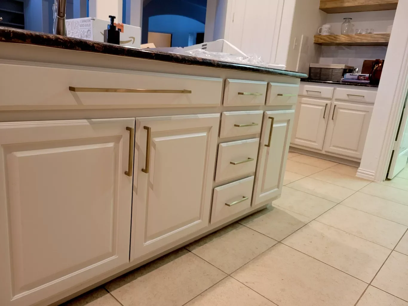 Kitchen with cream-colored cabinets with gold handles, a marble countertop, and tiled floor. Shelves with cups and glasses are visible in the background.