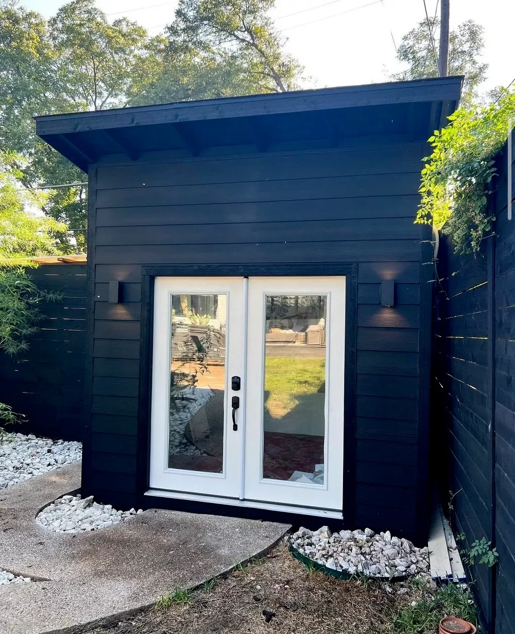 A small black shed with a sloped roof and double glass doors, surrounded by a landscaped yard with white rocks and a concrete pathway.