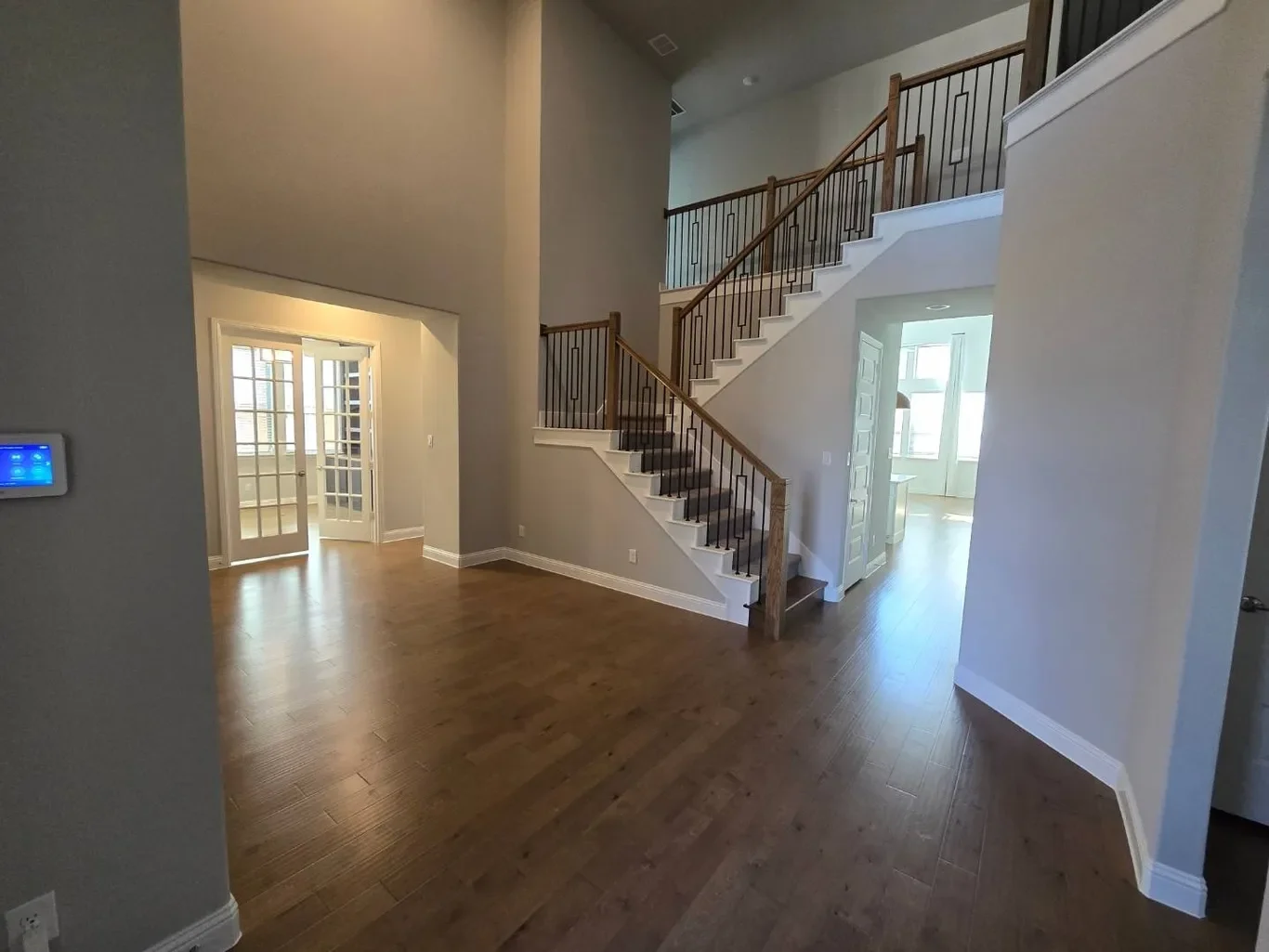 Interior view of a modern house showing a staircase with wooden railing, hardwood flooring, and doorway leading to a room with large windows.