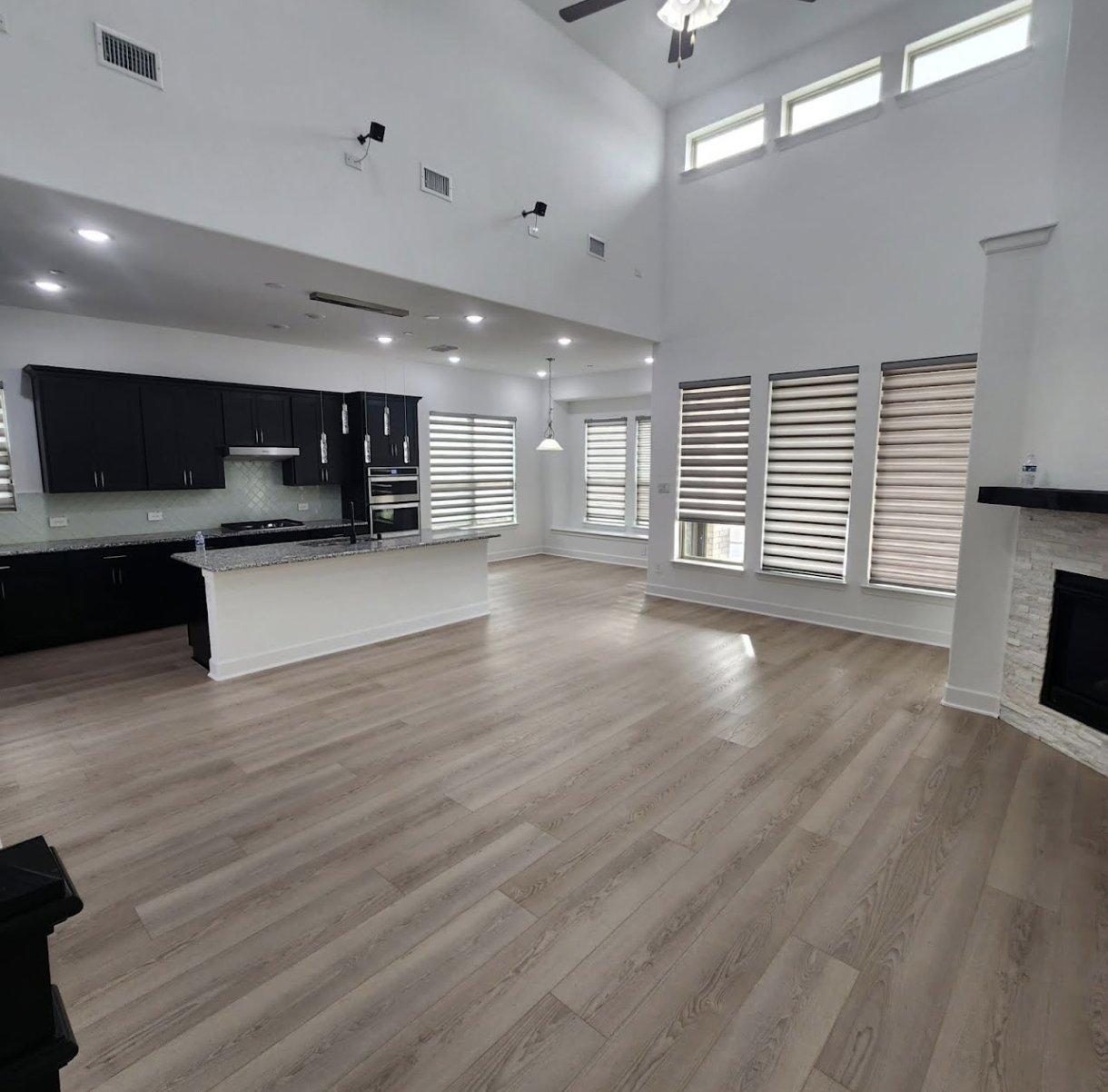 Open-plan living room and kitchen with light wood flooring, white walls, and multiple windows with blinds, featuring black kitchen cabinets and a fireplace.