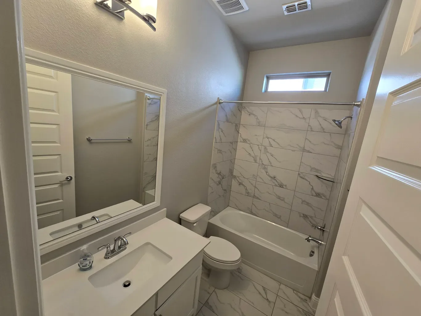 A small bathroom with a white vanity and sink, a mirror, toilet, bathtub with marble tile surround, small window above, and light-colored walls.