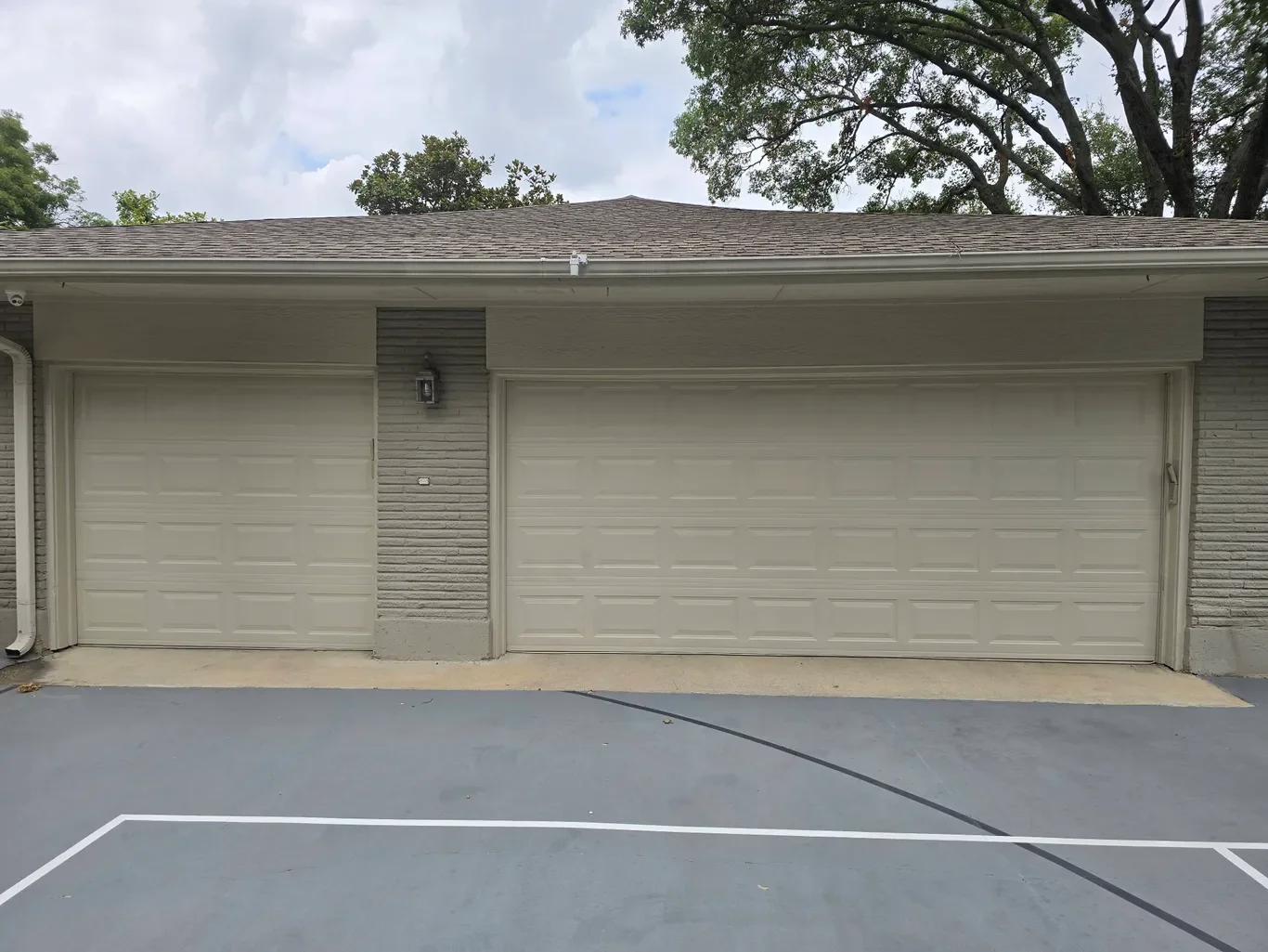 View of a residential garage with two white garage doors, gray brick accents, and a gray concrete driveway. Trees and cloudy sky in background.