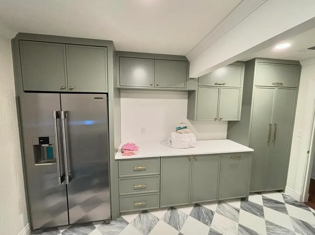 A kitchen with gray cabinets, a stainless steel refrigerator, white countertop, and a black and white diamond pattern tiled floor. There are some folded clothes and papers on the countertop.