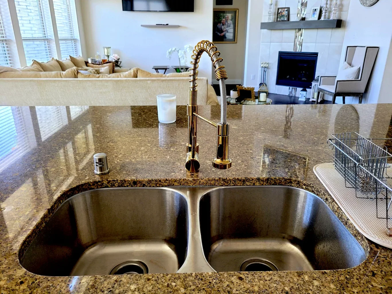 Close-up of a kitchen sink with a modern gold faucet, brown granite countertop, and a dish rack on the right. The background shows a living room with a beige sofa, windows with blinds, and a fireplace with a chair nearby.