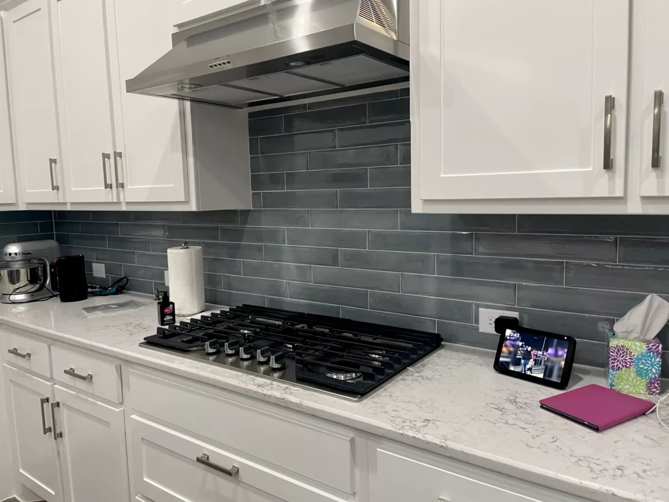 Kitchen countertop with white cabinets, gray backsplash, stove, coffee maker, paper towel roll, tablet, pink notebook, tissue box, and a mixer.