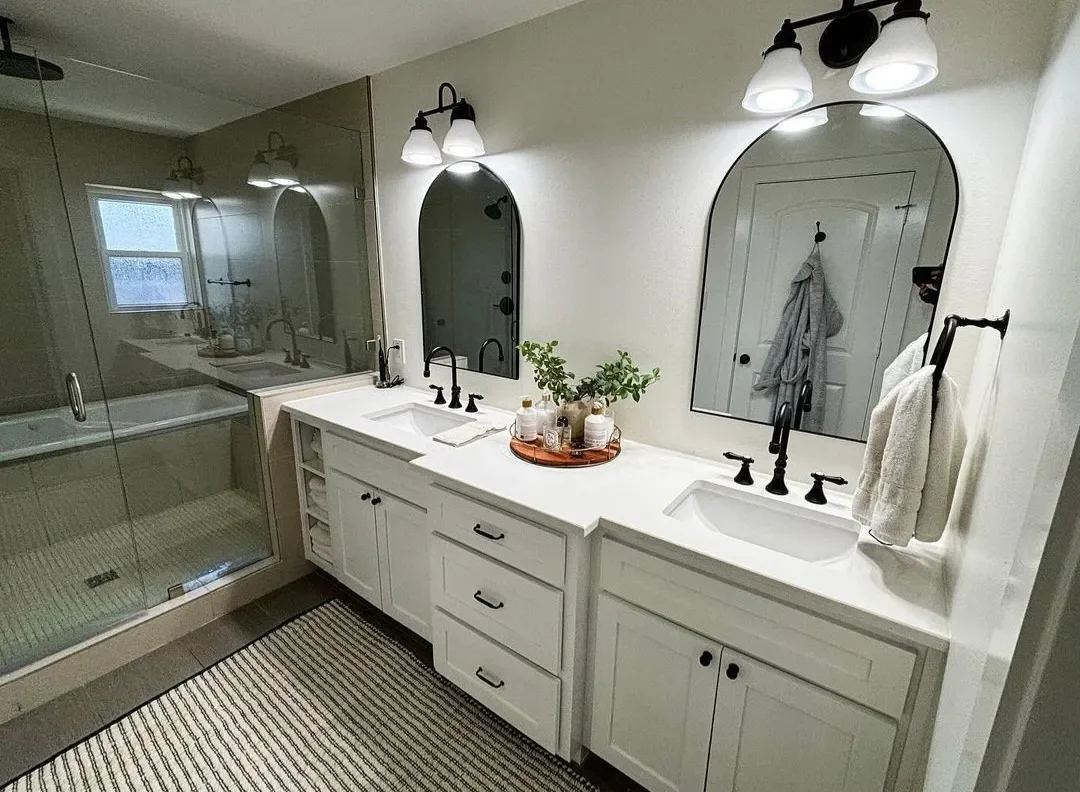 Bathroom with double vanity sinks and mirrors, black fixtures, wall-mounted lights, a towel hanging on a black towel holder, a decorative tray with toiletries and greenery, a glass-enclosed shower, a window, and a striped rug on the floor.