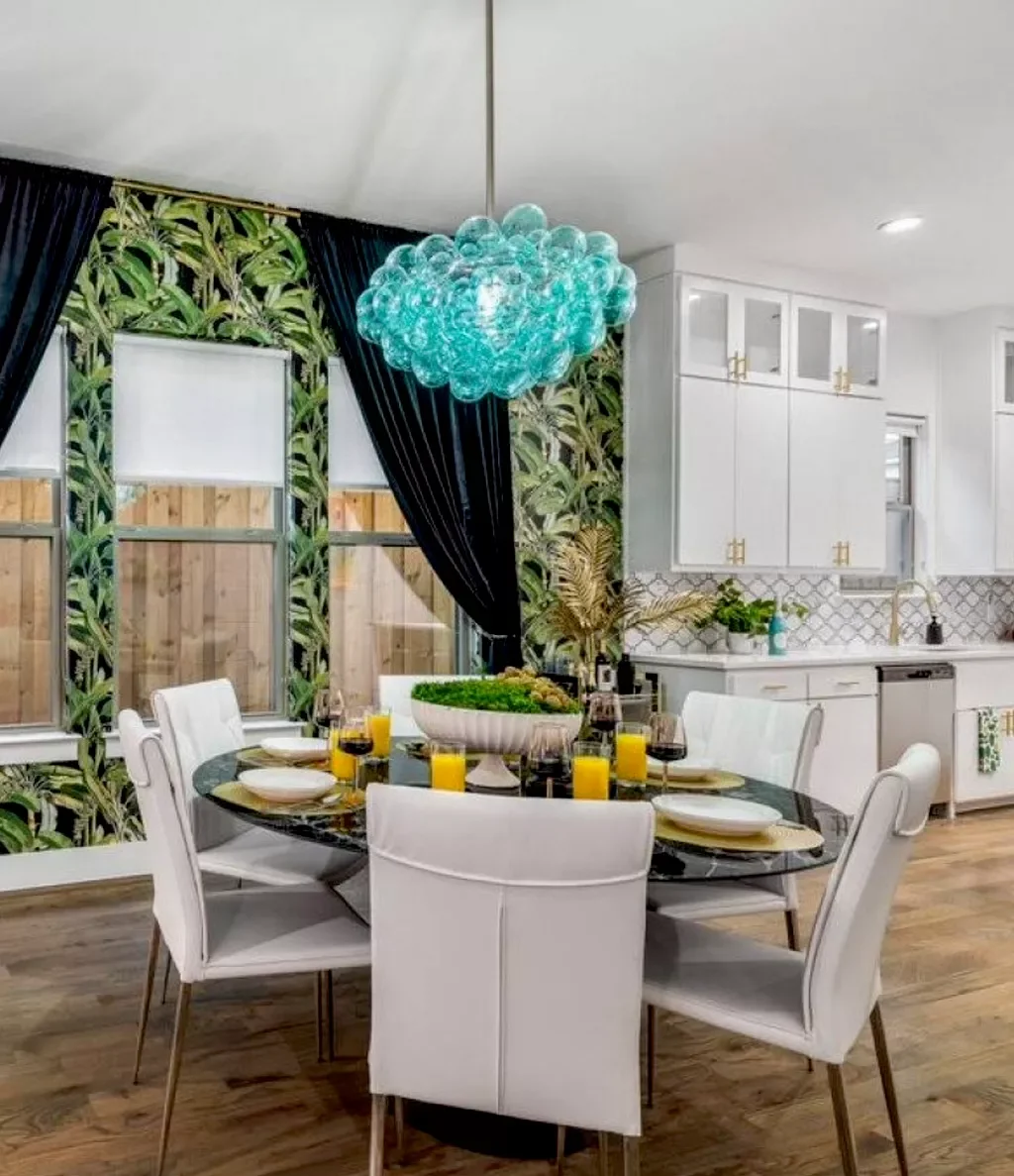 Dining room with a round table set with white plates, yellow drinks, wine glasses, and a green centerpiece. There are six white chairs around the table. The room has a tropical wallpaper on one wall, large windows with black curtains, and a modern ch