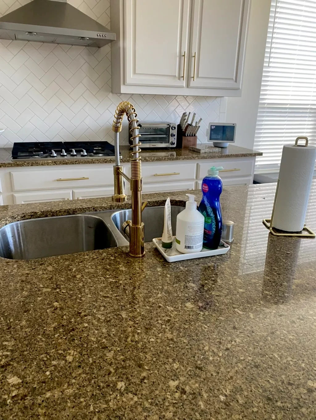 Kitchen countertop with a sink, gold faucet, cleaning supplies, paper towels, and small kitchen appliances, with cabinetry and blinds in the background.