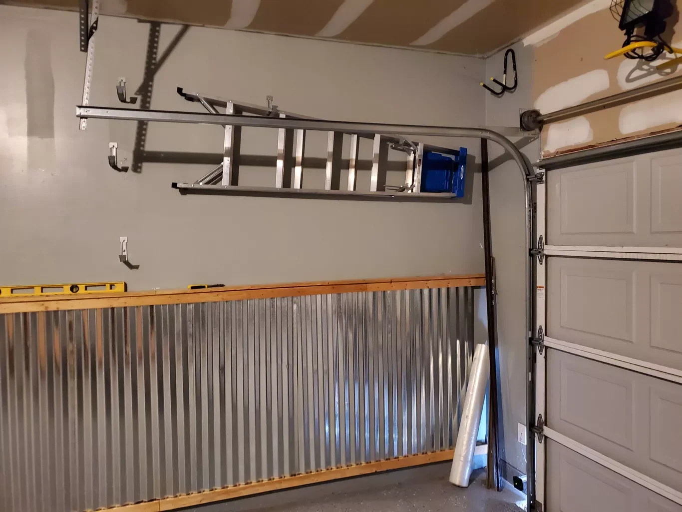 A garage with a metal ladder, a yellow level, hooks on the wall, and construction materials including corrugated metal and a rolled-up pipe leaning against the garage door.