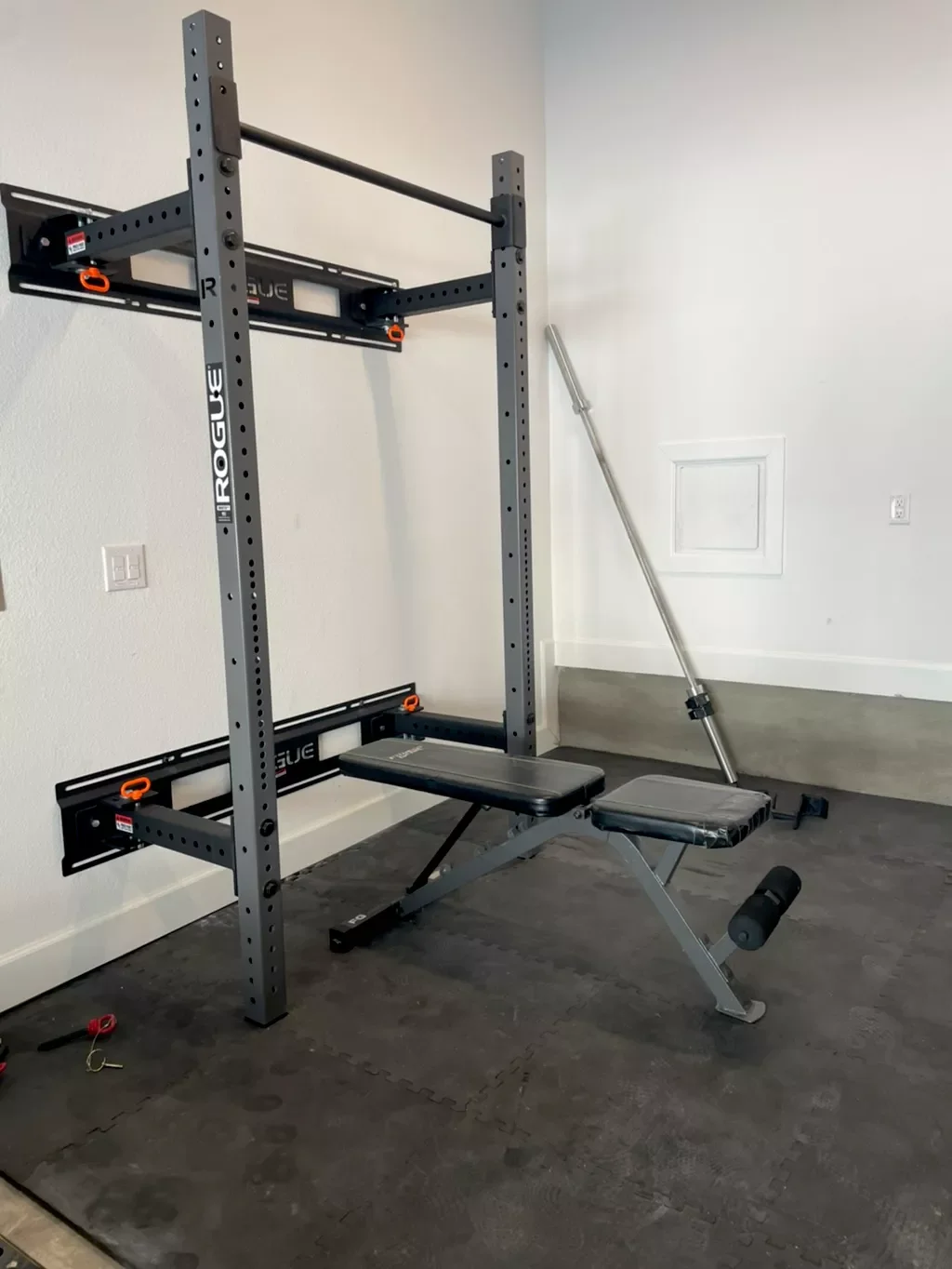 A home gym with a black and gray Rogue squat rack, a black adjustable workout bench, and a metal barbell on the wall. The floor is covered with black interlocking foam mats.