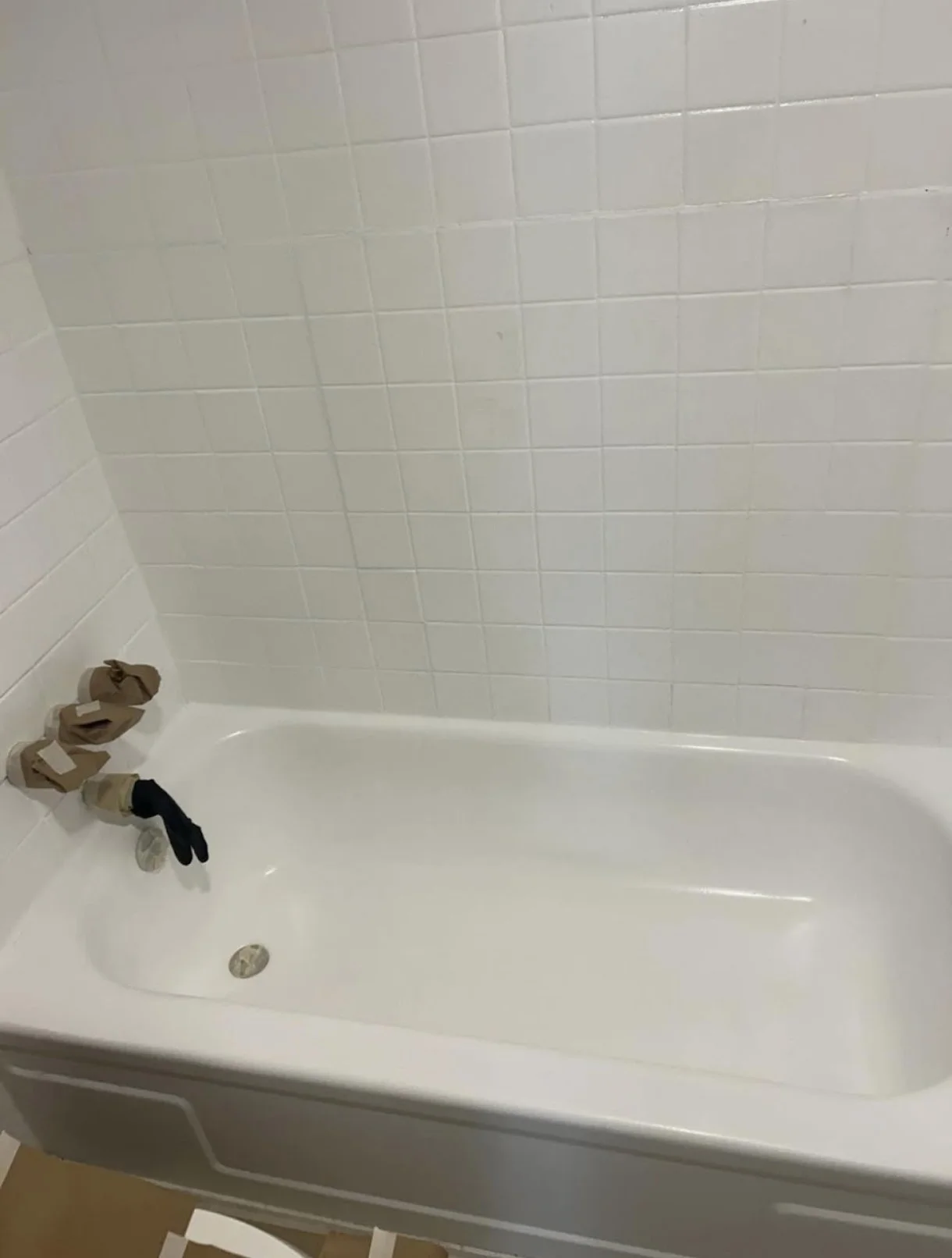 Empty white bathtub with a drain at the bottom, against a wall with white tiled tiles. There are two brown paper bags and a black glove resting on the edge of the bathtub.