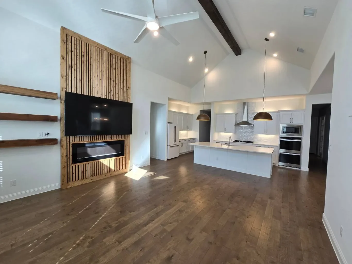Spacious open-plan kitchen and living area with white walls, hardwood floors, a large flat-screen TV mounted on a wooden slatted wall, a modern fireplace below, and kitchen cabinets, island with pendant lights, and stainless steel appliances.