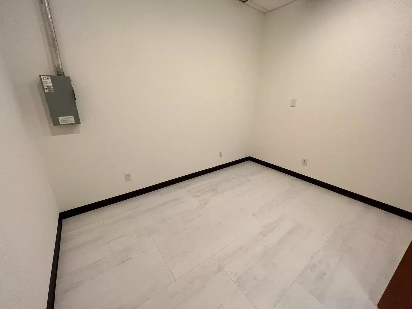 Empty room with beige walls, black baseboards, and a white tiled floor. An electrical panel is mounted on the left wall near the corner.