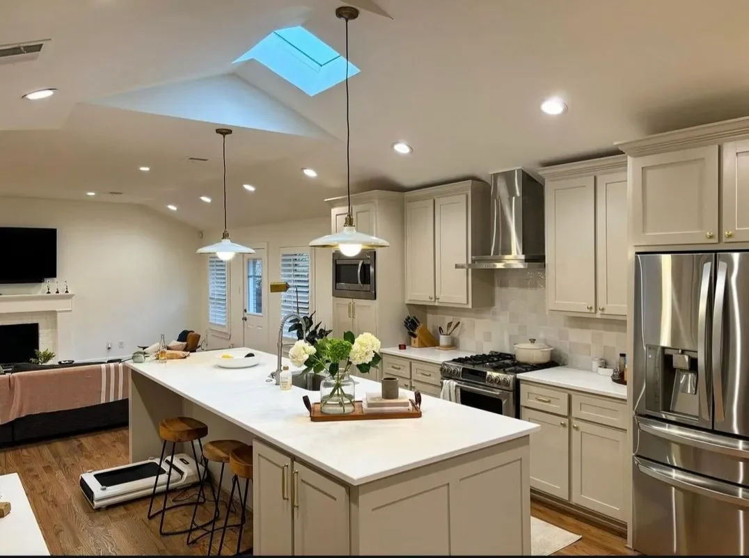 Modern kitchen with white cabinets, stainless steel appliances, a kitchen island with flowers, pendant lighting, a skylight, and bright recessed lighting, opening to a living area with a fireplace and a flat-screen TV.