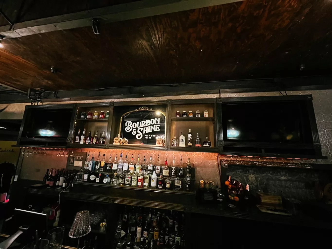 Bar with liquor bottles, shot glasses, and a illuminated sign that reads 'Bourbon & Shine' in a dimly lit setting.