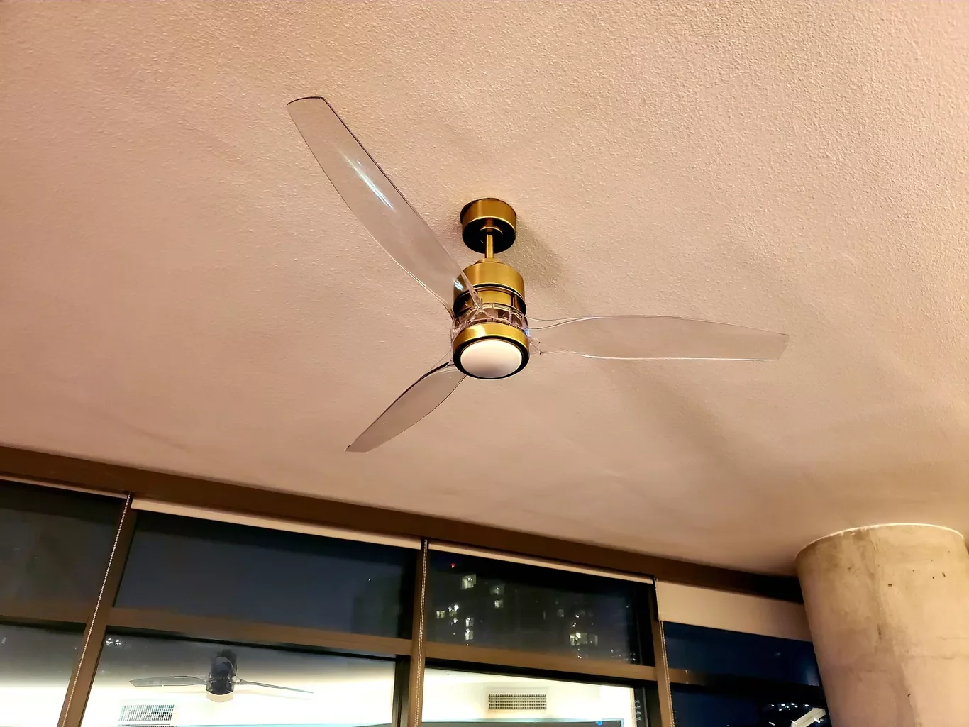 Ceiling fan with three transparent blades and a round light fixture in the center, mounted on a beige textured ceiling, with a glass window or door below showing indoor lighting and reflections.