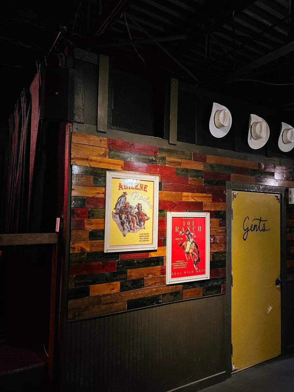 A wall with two vintage posters featuring rodeo and cowboy themes, a yellow door labeled 'Gents', and a wooden accent wall in a dimly lit space.