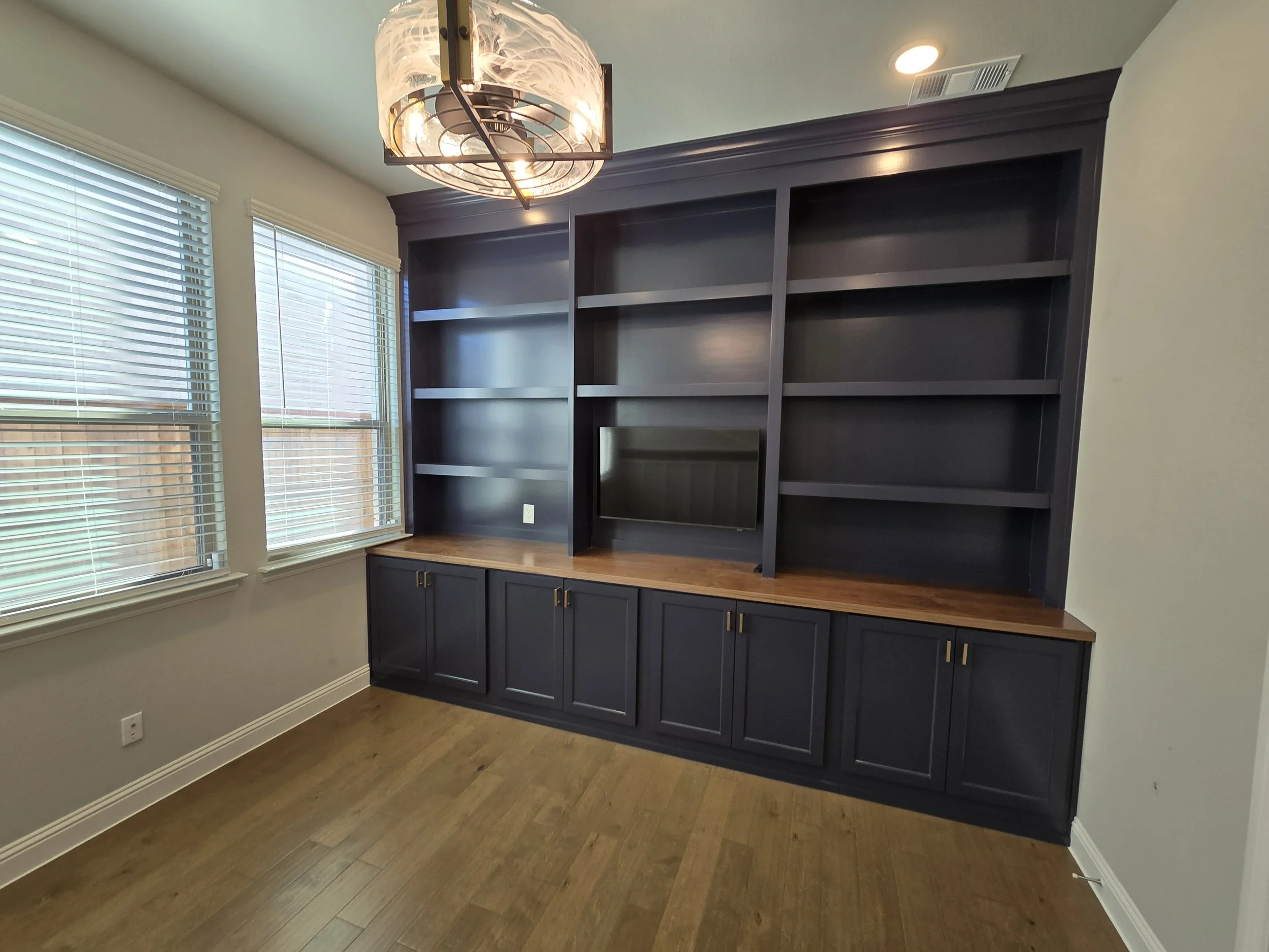 Empty built-in black bookshelf with a TV in the middle, wooden countertop, and cabinet doors below, in a room with windows and wooden floor.