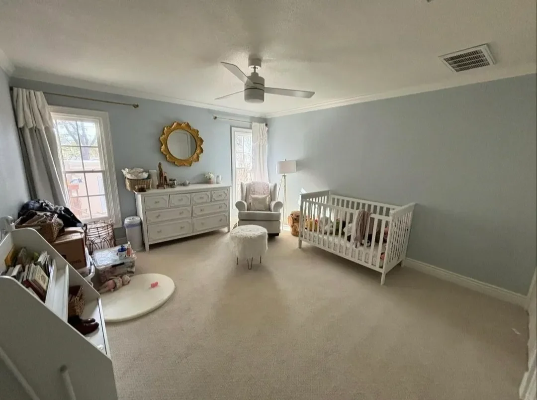 A nursery room with two windows, a white dresser, a white armchair, a white crib with toys, a standing lamp, and various small furniture and items, with light blue walls and beige carpet.