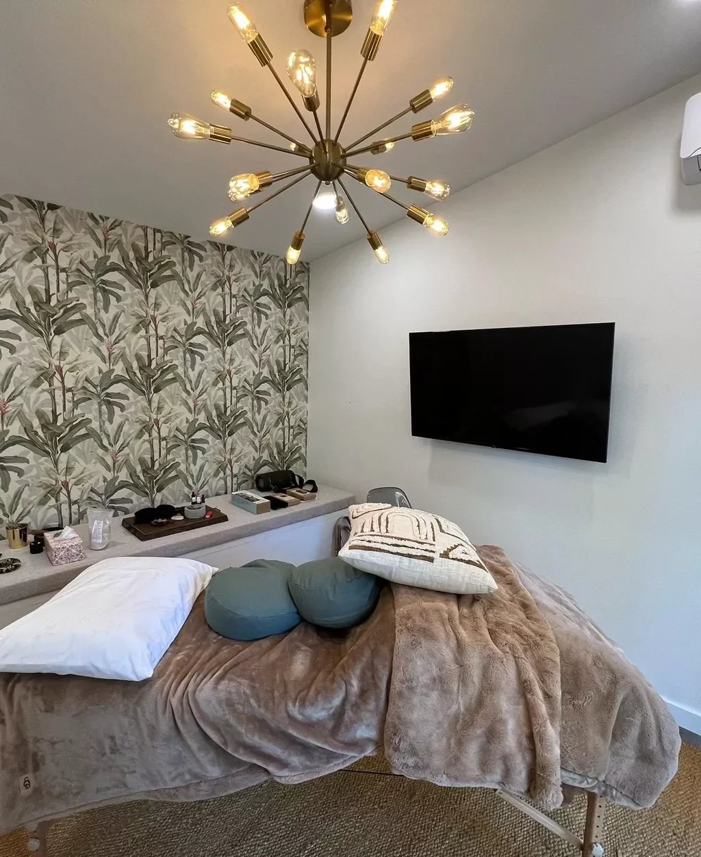 A bedroom with a bed covered in a brown plush blanket, pillows, and decorative cushions. There is a wall-mounted flat-screen TV and a decorative wallpapered wall with a botanical pattern. A modern chandelier with multiple exposed bulbs hangs from the
