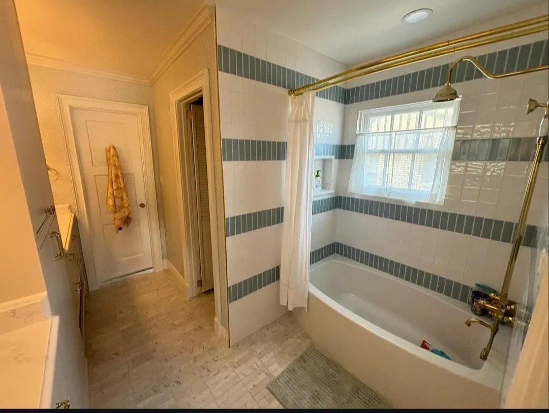 Bathroom with a white bathtub, gold fixtures, window with white curtain, tiled walls with blue horizontal stripes, and a window seat.