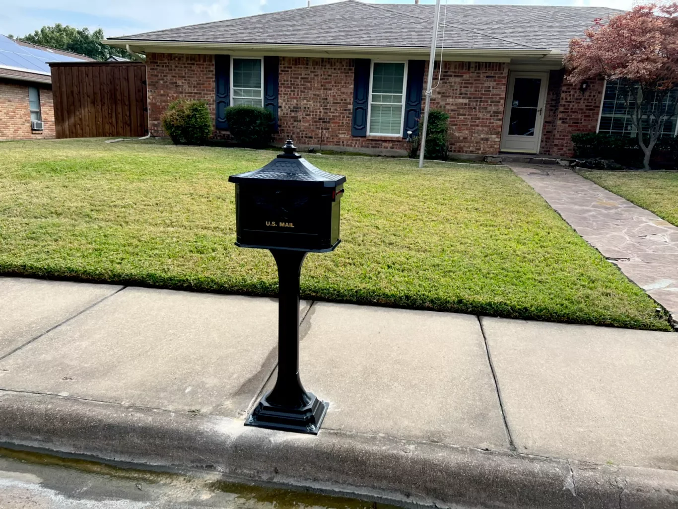 A black mailbox labeled 'U.S. Mail' on a post in front of a brick house with a manicured lawn, stone walkway, and trees.