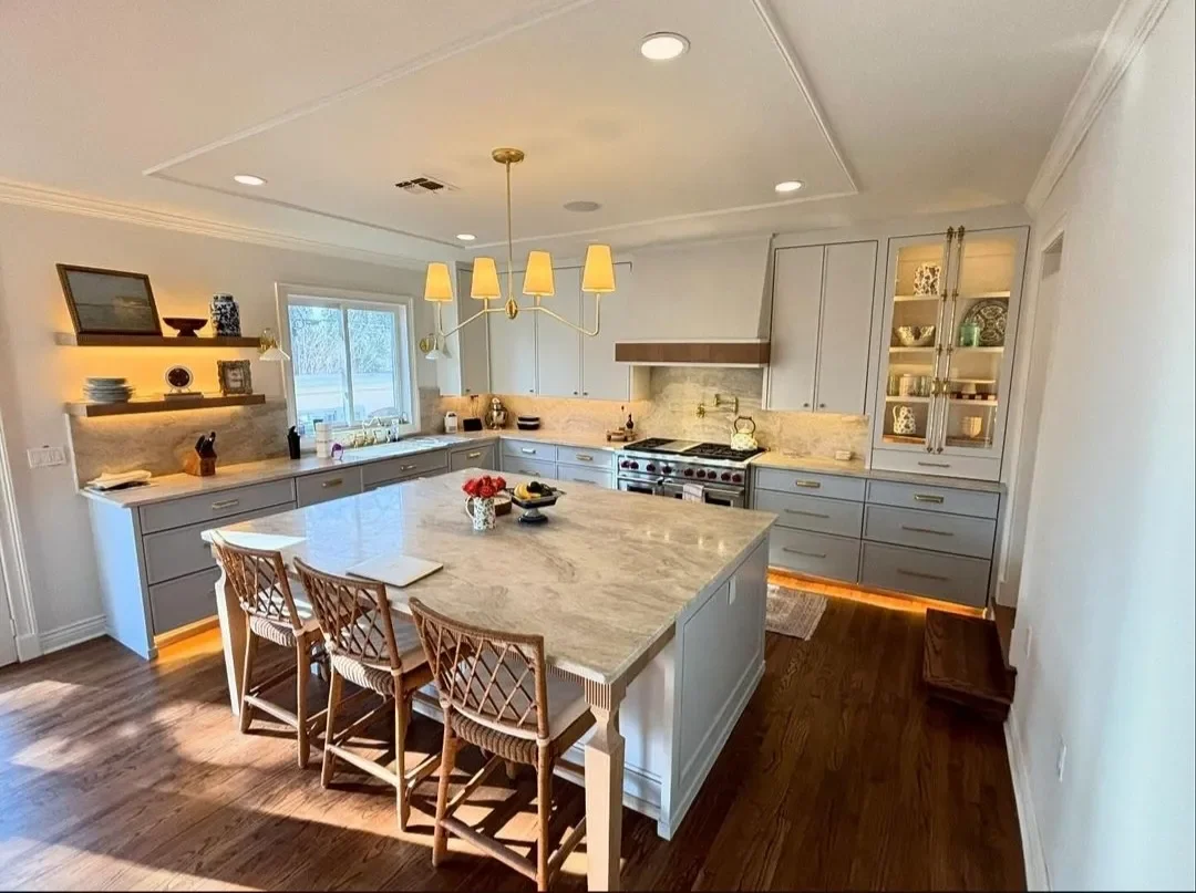 Modern kitchen with light gray cabinets, a large marble island, wooden flooring, and a chandelier above the island. There is a window with natural light, and open shelves with dishes and decorative items.