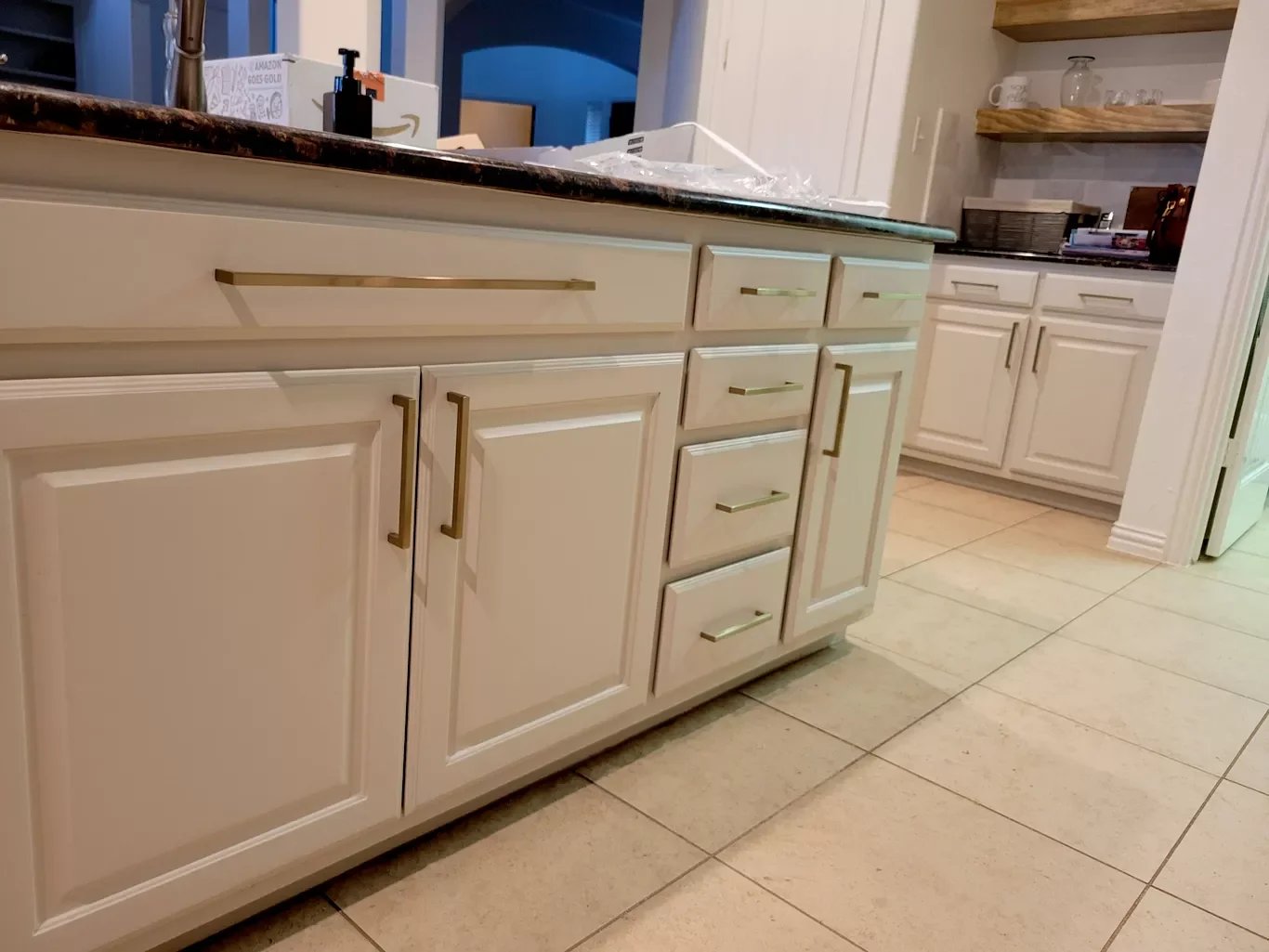 Kitchen with cream-colored cabinets with gold handles, a marble countertop, and tiled floor. Shelves with cups and glasses are visible in the background.